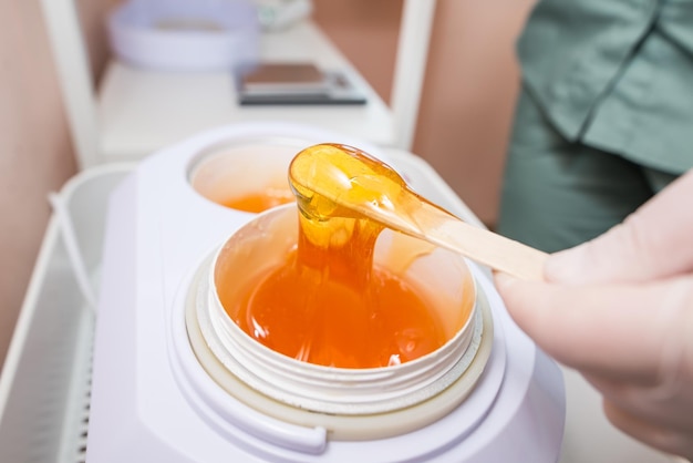 Pâte à sucre ou miel de cire dans un pot blanc Shugaring Concept d'épilation et de beauté La main de l'esthéticienne ramasse la pâte à sucre avec un bâton en bois