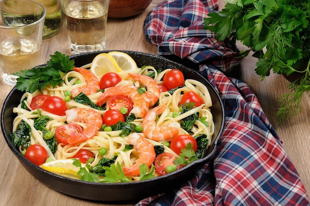 Pâtes aux crevettes frites, petits pois, tomates et épinards dans une poêle à frire, sur une table avec des verres à cidre.