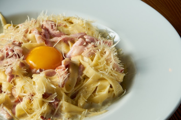 Pâtes Carbonara italiennes fraîches faites maison avec jaune d'oeuf cru, parmesan, sauce blanche, bacon dans un bol blanc sur fond de bois.