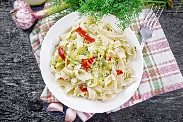 Pâtes fettuccine aux courgettes et piment rouge dans une sauce crémeuse dans une assiette blanche sur une serviette, de l'ail et une fourchette sur fond de planche de bois d'en haut