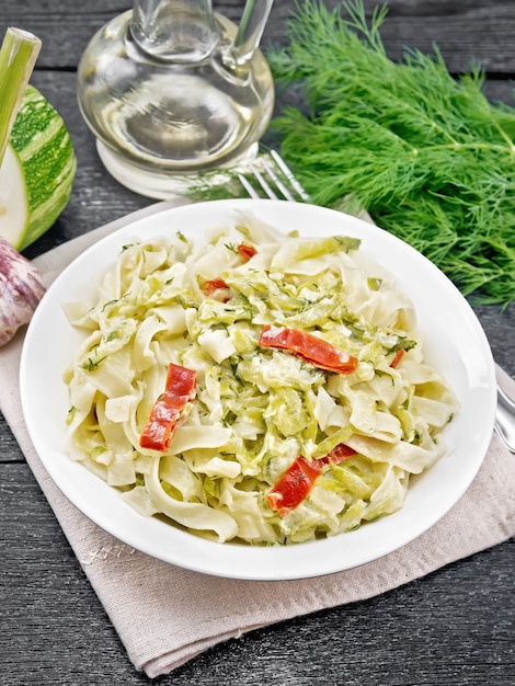 Pâtes fettuccine aux courgettes et piment rouge dans une sauce crémeuse dans une assiette blanche sur une serviette, de l'ail et une fourchette sur fond de planche de bois