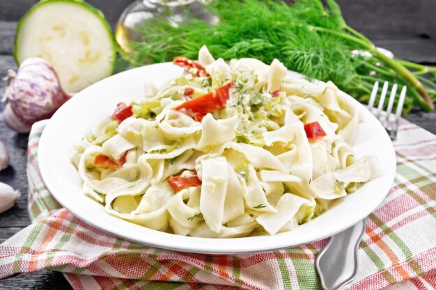 Pâtes fettuccine aux courgettes et piment rouge dans une sauce crémeuse dans une assiette blanche sur un torchon, de l'ail et une fourchette sur fond de planche de bois foncé