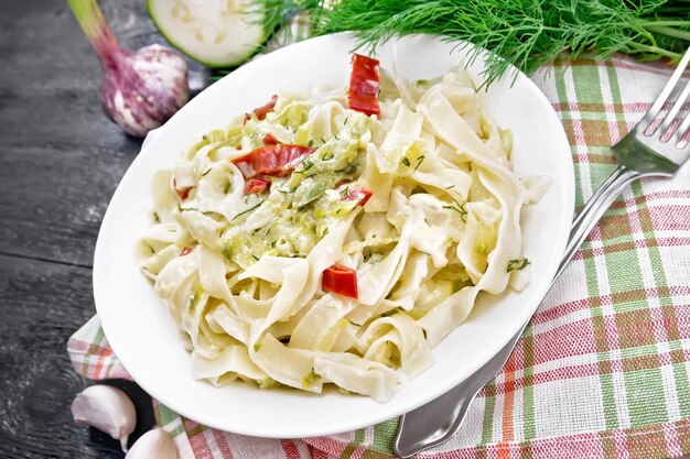 Pâtes fettuccine aux courgettes et piments rouges dans une sauce crémeuse dans une assiette blanche sur une serviette à l'ail et une fourchette sur fond de planche de bois