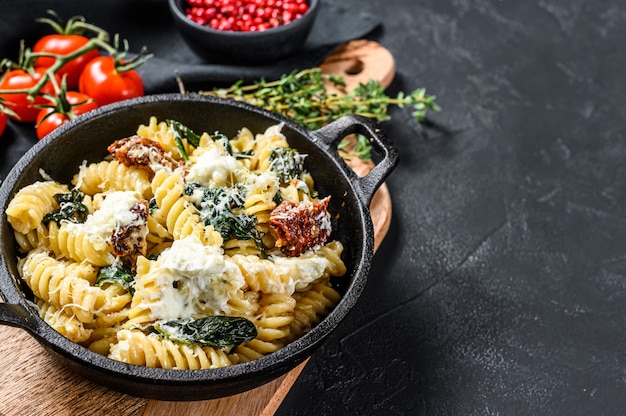 Pâtes fusilli aux épinards, tomates séchées et ricotta dans une poêle. Fond noir. Vue de dessus. Espace copie