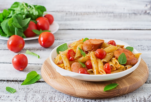 Pâtes penne à la sauce tomate avec saucisse, tomates, basilic vert décorées dans une poêle sur une table en bois