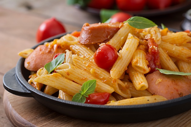 Pâtes penne à la sauce tomate avec saucisse, tomates, basilic vert décorées dans une poêle sur une table en bois