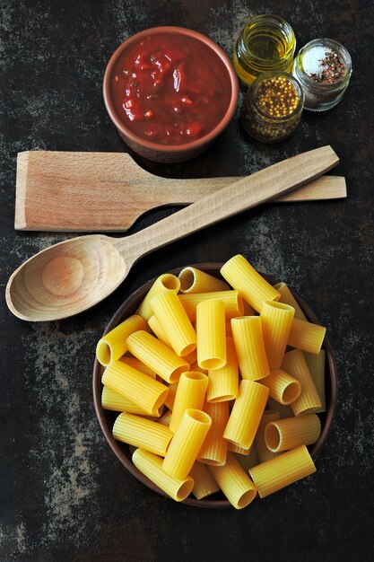 Pâtes rigatoni non cuites dans un bol. Sauce tomate et épices. Spatule et cuillère en bois. Concept de cuisson des pâtes à la sauce tomate. Vue de dessus. Mise à plat.