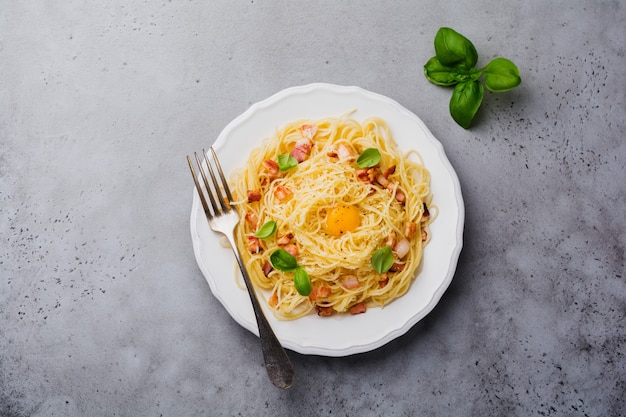 Pâtes spaghetti carbonara avec bacon, parmesan, jaune d'oeuf et feuilles de basilic sur une surface grise claire. Plat italien traditionnel