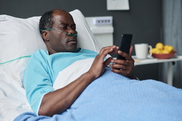 Photo patient âgé afro-américain utilisant un smartphone allongé sur son lit à l'hôpital