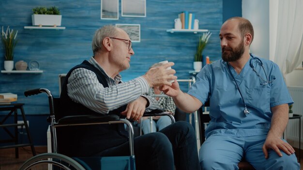 Patient âgé Atteint D'une Maladie Chronique Recevant Une Bouteille De Pilules Comme Traitement D'une Infirmière Dans Une Maison De Soins Infirmiers. Vieil Homme En Fauteuil Roulant Parlant à Un Assistant Médical De Médicaments Et De Prescription