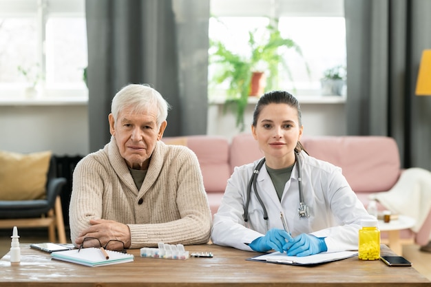 Patient âgé malade en tenue décontractée et jeune femme médecin en blouse blanche et gants assis l'un à côté de l'autre et vous regardant