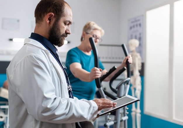 Patient âgé utilisant un vélo stationnaire pour la récupération physique pendant que le médecin regarde une tablette pour les soins de santé. Femme blessée faisant de l'exercice pour le fitness et la physiothérapie