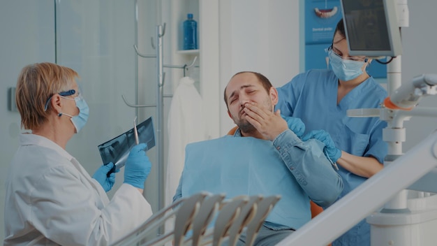 Patient se plaignant de maux de dents lors d'une consultation de stomatologie pendant que le dentiste analyse la radiographie dentaire pour effectuer une procédure de forage. Stomatologue principal traitant les caries pour les soins bucco-dentaires.