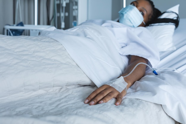 Photo une patiente afro-américaine portant un masque, avec un goutte-à-goutte à la main, allongée sur un lit dans une chambre d'hôpital.