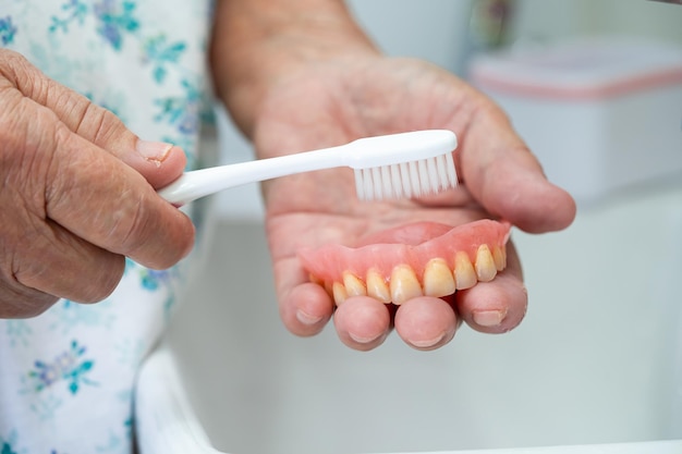 Une patiente asiatique âgée ou âgée utilise une brosse à dents pour nettoyer la prothèse partielle des dents de remplacement