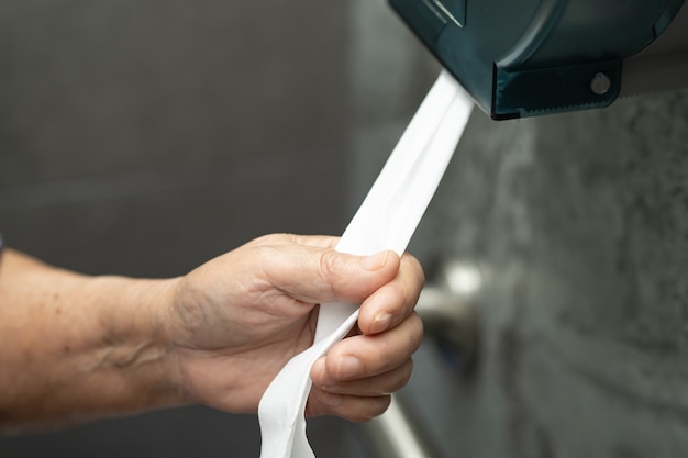 Une patiente asiatique âgée tire le mouchoir du rouleau dans la salle de bain des toilettes