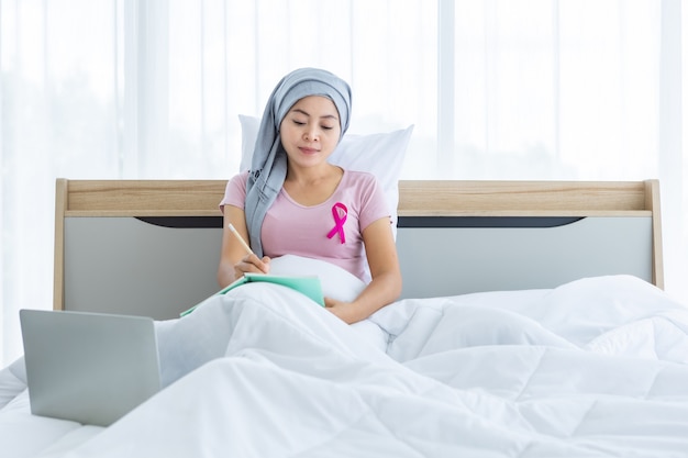 Une patiente asiatique atteinte d'un cancer du sein avec un ruban rose portant un foulard Après un traitement à la chimiothérapie avec Prendre des notes dans un cahier et un ordinateur portable sur le lit Dans la chambre à la maison, soins de santé