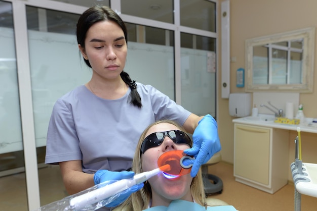 Patiente portant des lunettes de protection allongée sur une chaise dans le bureau de la clinique dentaire Dentiste mettant un remplissage de photopolymère à l'aide d'un équipement moderne