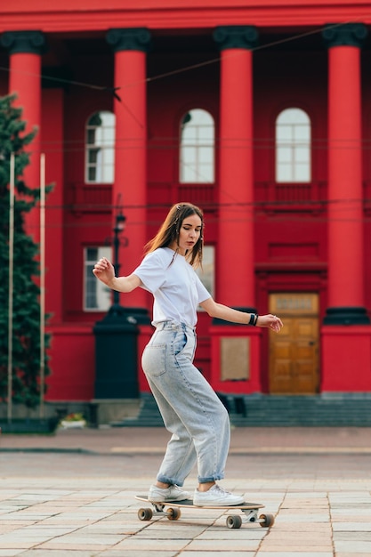 Patineuse en vêtements amples sur sa longue planche dans une rue de la ville devant un bâtiment rouge