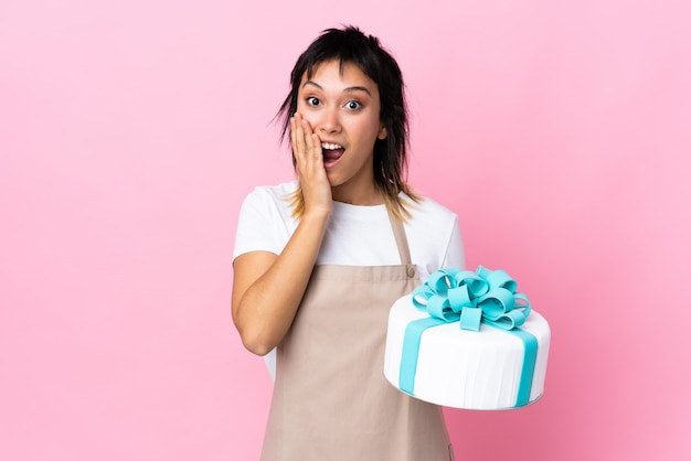 Pâtissier uruguayen tenant un gros gâteau sur rose isolé avec une expression faciale surprise