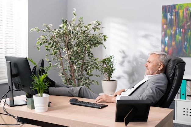 Un patron d'entreprise âgé souriant et détendu est assis dans une chaise confortable dans une position détendue avec les pieds