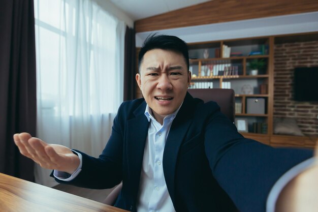 Le patron d'un homme d'affaires asiatique en colère parle par appel vidéo à l'aide d'un téléphone travaillant dans un bureau classique se disputant avec un employé regardant la caméra du téléphone