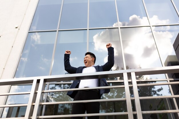 Le patron d'un homme d'affaires asiatique se tient près de son centre de bureau sur le balcon dans un costume de couleur sombre et tient les mains en l'air pour célébrer la victoire