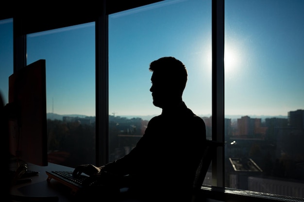 Patron d'homme d'affaires prospère travaillant à l'ordinateur sur le fond de grandes fenêtres, silhouette