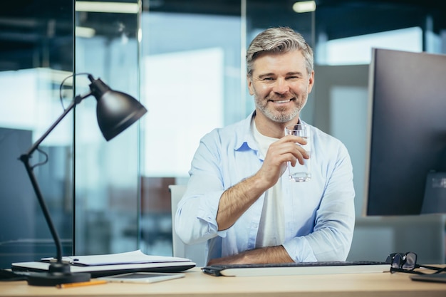 Patron d'homme d'affaires de travailleur prospère regardant la caméra et souriant tenant un verre d'eau