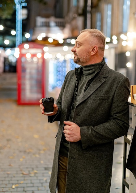 Pause café. Homme mûr en manteau élégant souriant gentiment tout en prenant un café. Rue du soir.