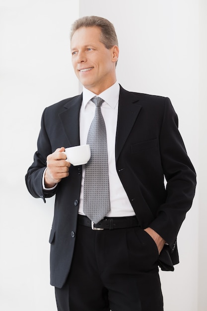 Pause café. Homme senior gai en tenue de soirée tenant une tasse de café et souriant tout en se tenant près de la fenêtre