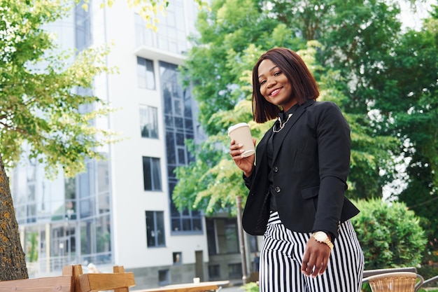 Pause-café Jeune femme afro-américaine dans des vêtements à la mode à l'extérieur de la ville près des arbres verts et contre le bâtiment de l'entreprise