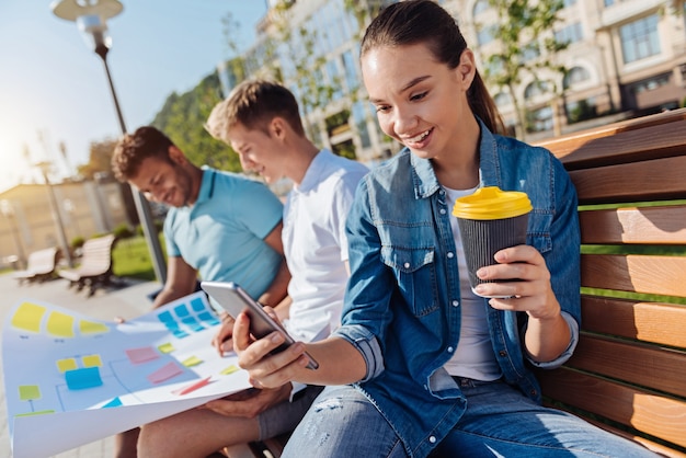 Pause café. Personne de sexe féminin ravi positif tenant une tasse de papier et en gardant le sourire sur son visage tout en regardant le téléphone