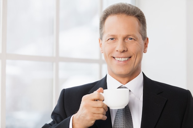 Pause café. Portrait d'un homme mûr gai en tenue de soirée tenant une tasse de café et vous souriant tout en se tenant près de la fenêtre