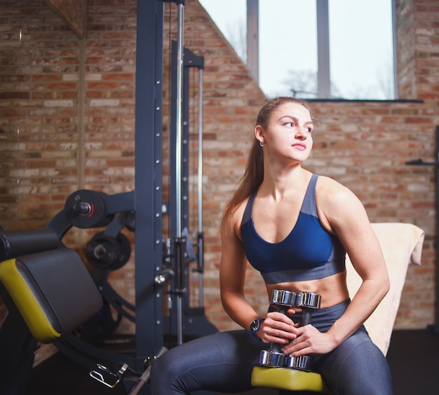 Pause d'entraînement. Jeune femme sportive en tenue de sport reposant sur un banc tenant des haltères dans ses mains et à l'écart dans la salle de gym