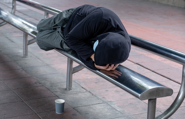 Un pauvre sans-abri dort sur une chaise d'arrêt de bus. Il y avait une tasse à mettre au mendiant parce que la pauvreté demandait de l'aide aux gens qui passaient dans les rues.