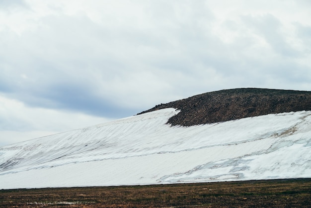 Paysage alpin minimaliste atmosphérique avec large glacier sur une pente de montagne rocheuse sous un ciel nuageux. Magnifique paysage de montagne avec flanc de montagne enneigé dans la vallée des hautes terres. Collines rocheuses avec de la neige.