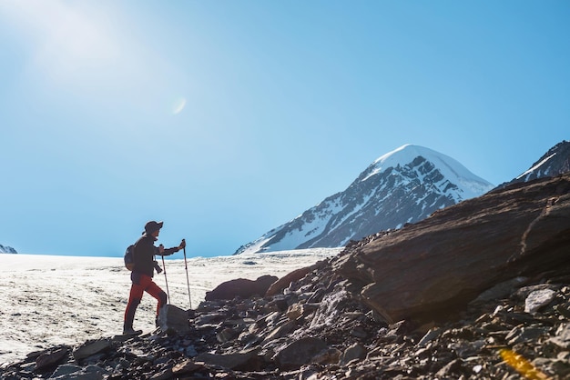 Paysage alpin pittoresque avec silhouette de randonneur avec des bâtons de randonnée contre une grande langue glaciaire et un sommet de montagne enneigé au soleil Homme avec sac à dos en haute montagne sous un ciel bleu par temps ensoleillé