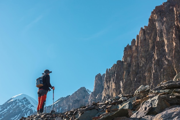 Paysage alpin pittoresque avec silhouette de randonneur avec des bâtons de trekking contre de gros rochers pointus et un sommet de montagne enneigé au soleil Homme avec sac à dos en haute montagne sous un ciel bleu par temps ensoleillé