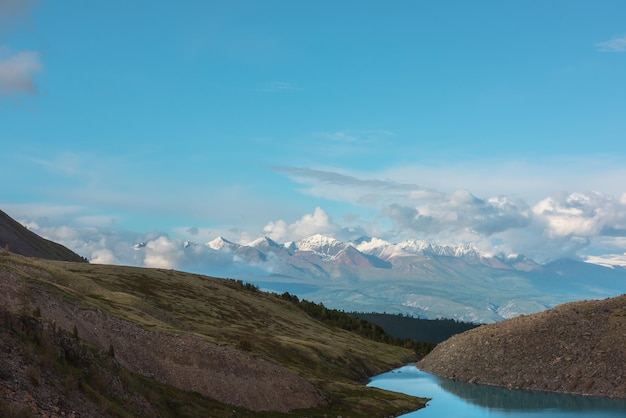 Paysage atmosphérique avec lac de montagne azur contre haute chaîne de montagnes blanches comme neige au soleil dans les nuages bas Vue spectaculaire en soirée sur le lac alpin et les grandes montagnes enneigées ensoleillées dans les nuages bas