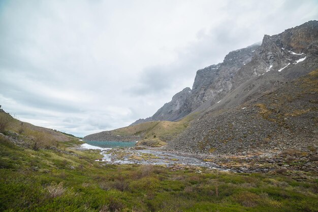 Paysage atmosphérique avec un lac de montagne azur dans une vallée verdoyante et une chaîne de montagnes rocheuses avec un sommet pointu dans des nuages bas Beau lac alpin et de hautes roches pointues sous un ciel nuageux par temps maussade