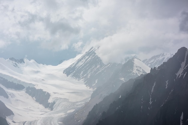 Paysage atmosphérique avec des silhouettes floues de rochers pointus et de sommet de montagne enneigé dans des nuages bas pendant la pluie Vue spectaculaire sur de grandes montagnes enneigées et un glacier flou dans la brume de pluie dans des nuages bas gris