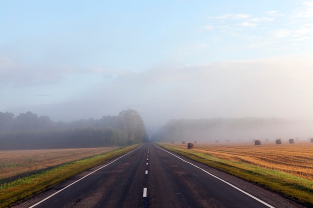 Paysage d'automne avec route et forêt pendant le brouillard, saison estivale, champ droit avec des piles de paille de céréales