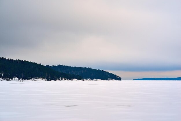 Paysage d'hiver avec ciel gris et neige blanche sur un grand lac et avec forêt sur un horizon vallonné