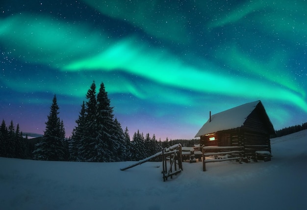 Paysage d'hiver fantastique avec maison en bois avec lumière dans la fenêtre dans les montagnes enneigées et lumière du nord dans le ciel nocturne Concept de vacances de Noël et de vacances d'hiver