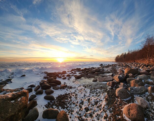 Paysage d'hiver Vue panoramique du magnifique coucher de soleil sur la baie Liste des nuages sur l'eau à la lumière brillante Neige glacée et rochers sur le littoral Le soleil couchant se reflète dans la mer