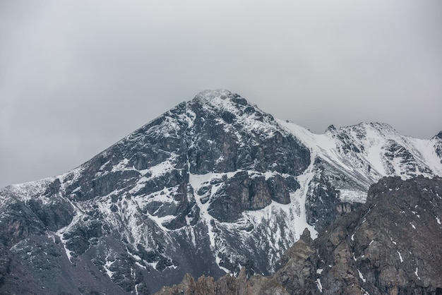 Photo paysage impressionnant avec une haute montagne enneigée au sommet avec des rochers pointus dans un ciel nuageux vue spectaculaire sur le pic pointu de la montagne enneigée par temps de pluie paysage atmosphérique avec de la neige blanche sur des rochers noirs