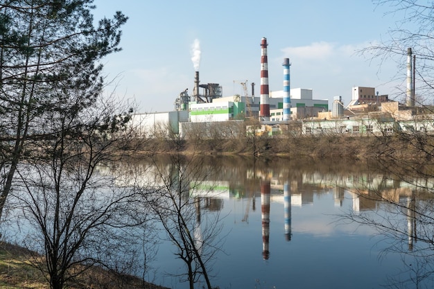 Paysage industriel Une grande usine avec des tuyaux se reflète dans l'eau claire de la rivière contre le ciel bleu De la fumée toxique provient de la cheminée de l'usine Pollution de l'environnement