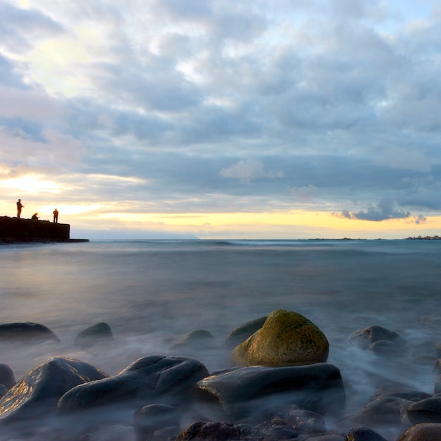 Paysage marin au coucher du soleil avec de l'eau de mer en flou de mouvement, des pierres sur la plage et des pêcheurs sur la jetée. Océan Atlantique, île de Ténérife. Longue exposition