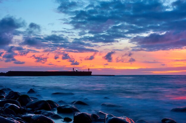 Paysage marin avec océan Atlantique et ciel au crépuscule, pierres sur la plage et pêcheurs sur la jetée. Longue exposition avec eau de mer floue
